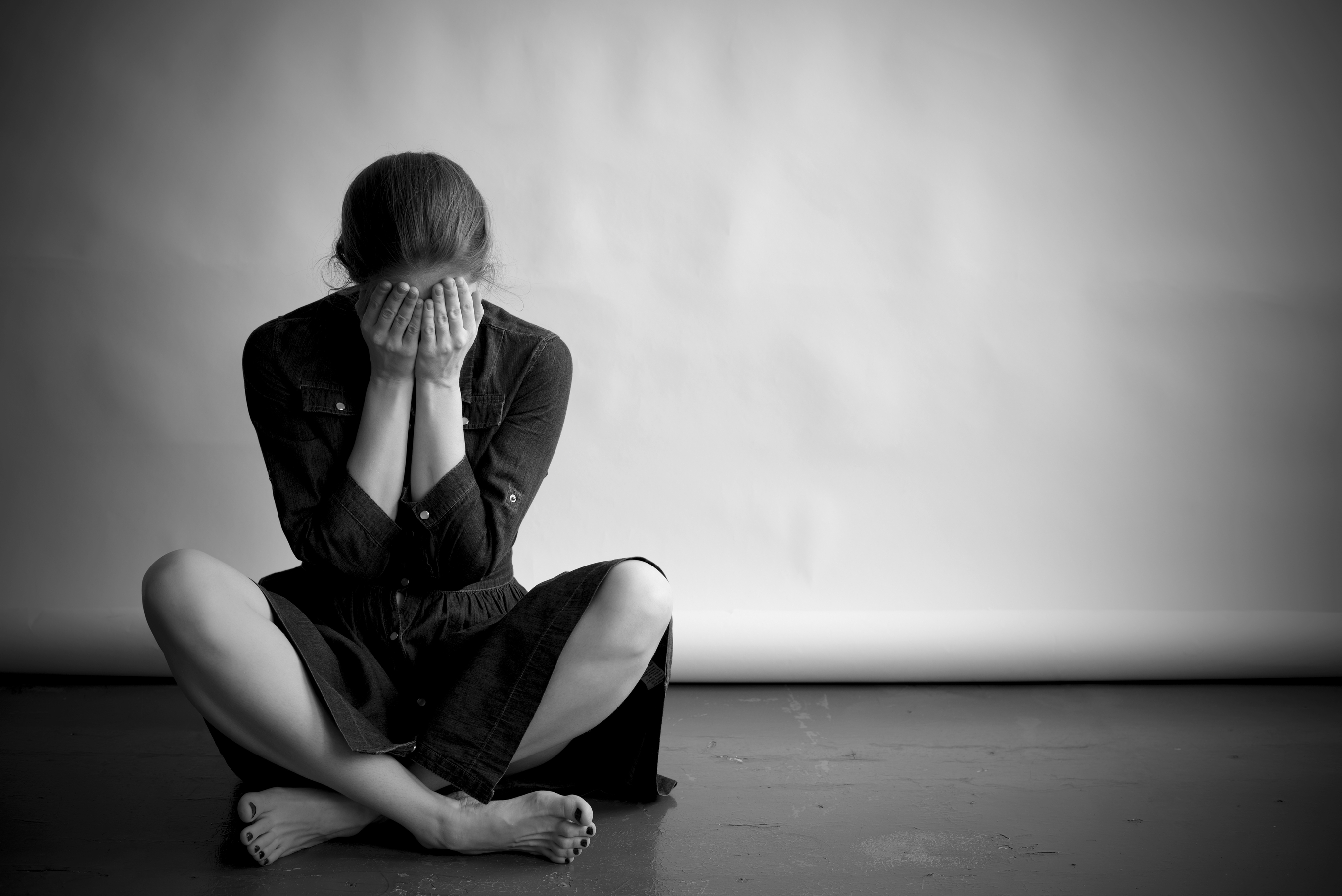 Woman is sitting on an old floor with cracks. She is sad and depressed, covering her face with hands. Studio paper background in behind her.